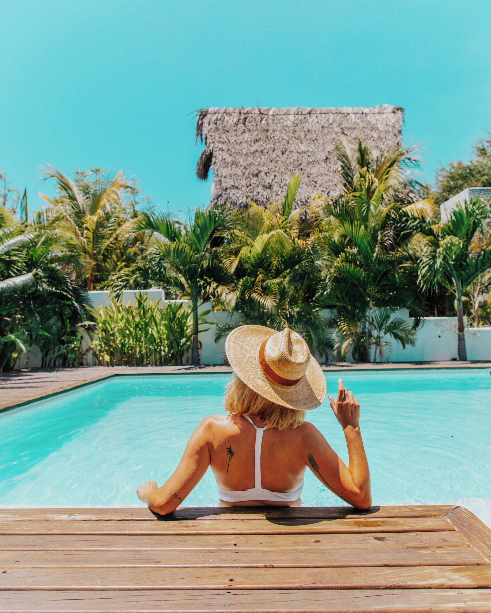 Eine Person entspannt am Pool mit Blick auf eine tropische Landschaft, darunter Palmen und ein Steinbauwerk.
