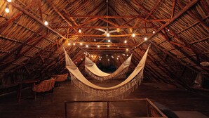 A thatched-roof room with a hammock, wooden furniture, and string lights.