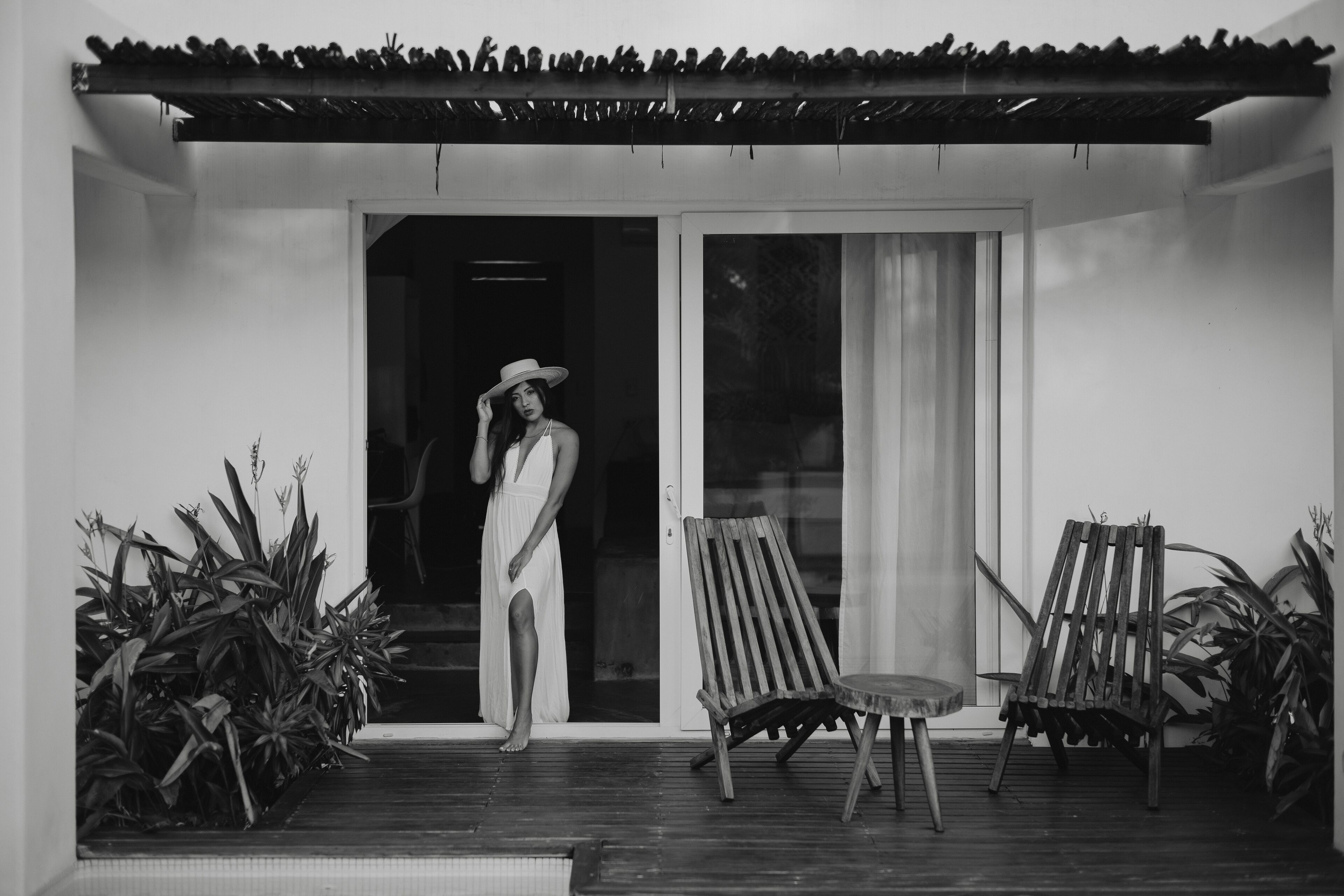A woman in a white dress standing on a wooden deck with two wooden chairs and a table.