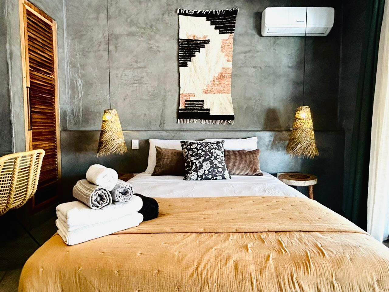 A bedroom with a large bed, a headboard, a wall-mounted air conditioner, and a woven wall hanging.