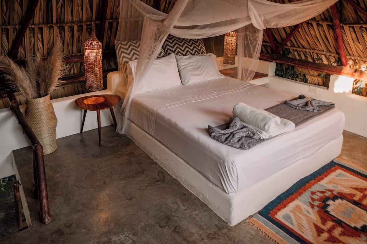 A room with a bed under a canopy, a small wooden table, and a woven lamp.