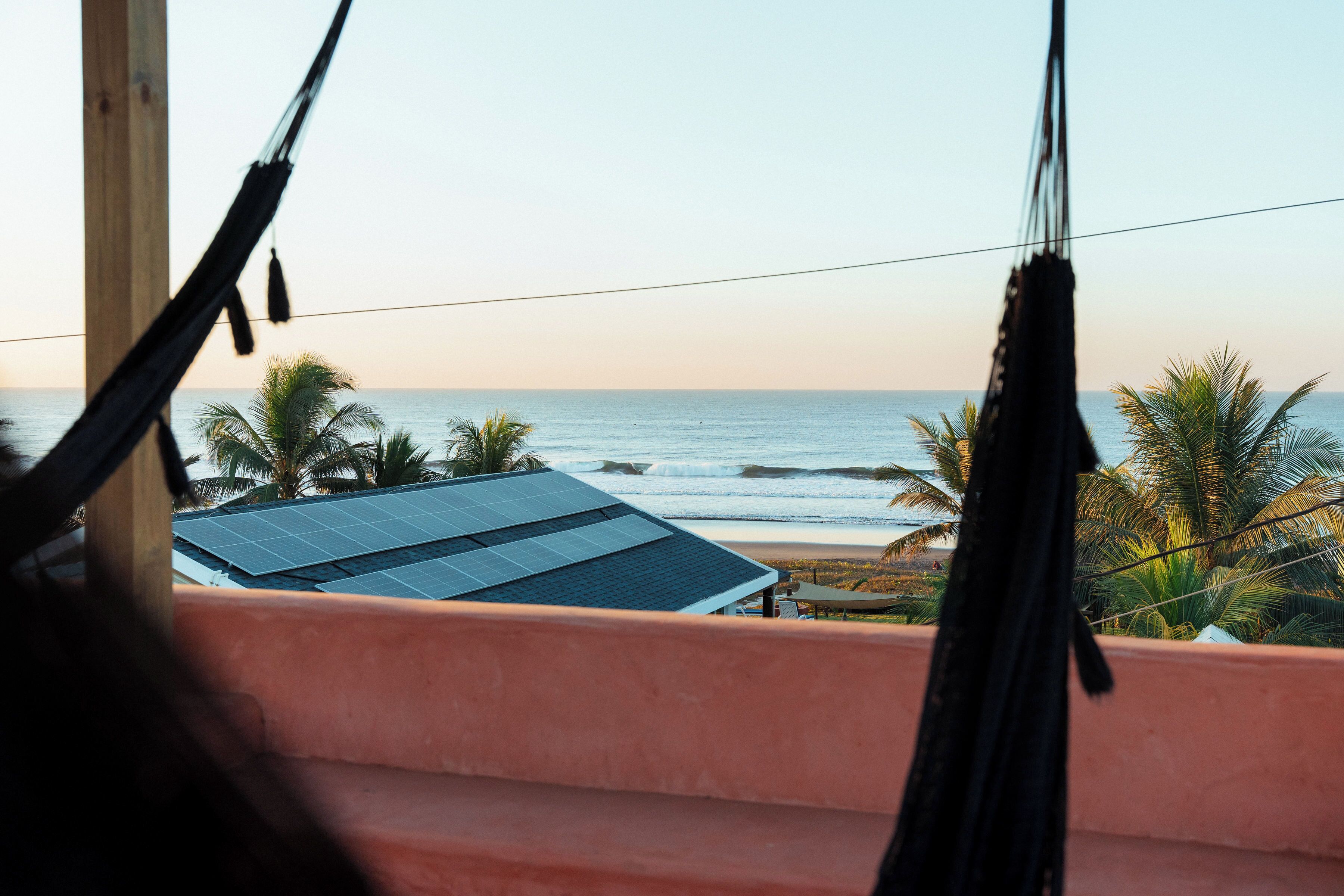 Un'amaca con vista su una spiaggia, palme e una casa con pannelli solari.