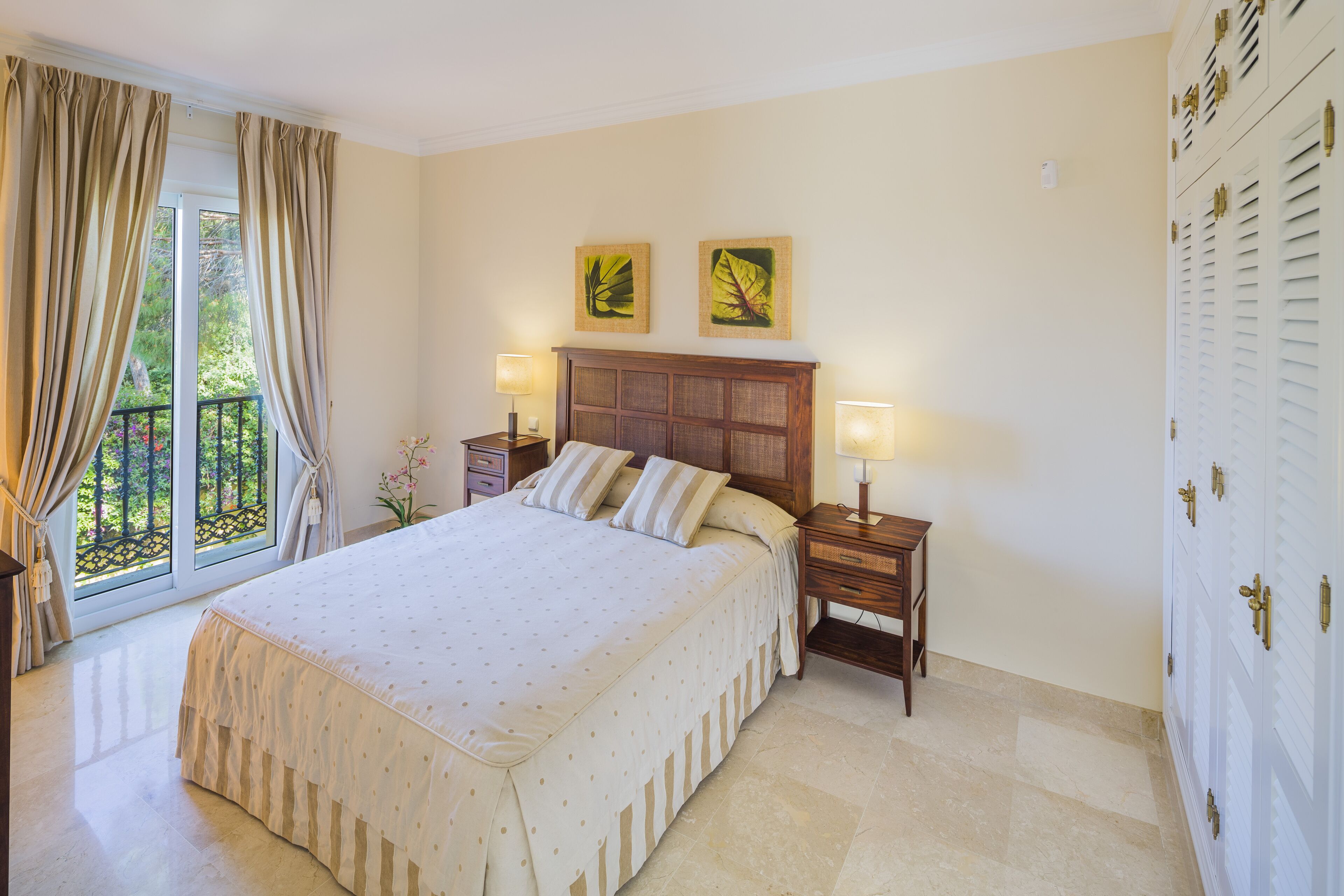 A bedroom with a large bed, two bedside tables with lamps, a wooden headboard, and a view of greenery through the window.