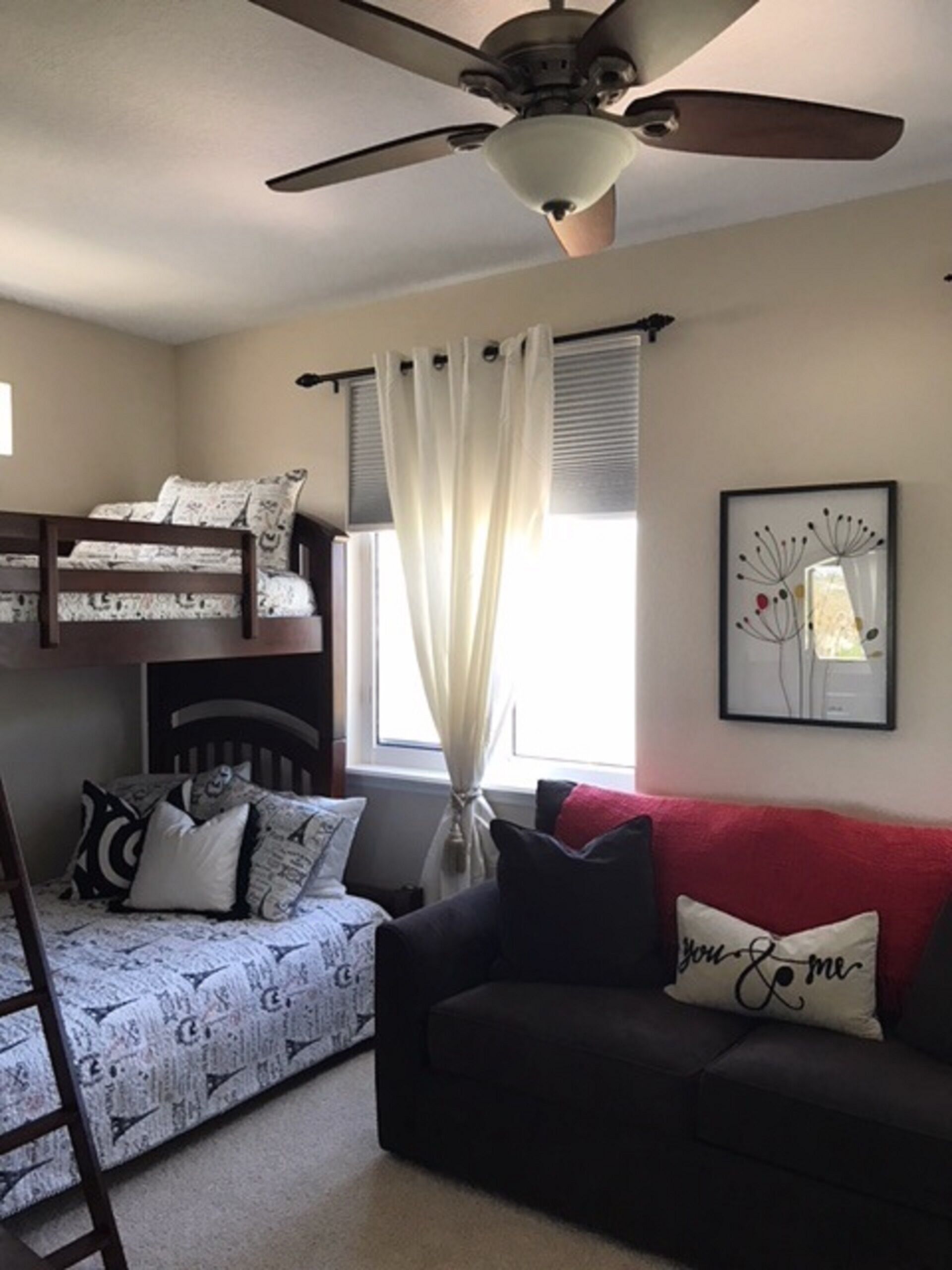 A bedroom with a bunk bed, a desk, a chair, a ceiling fan, a window with curtains, and a framed picture on the wall.