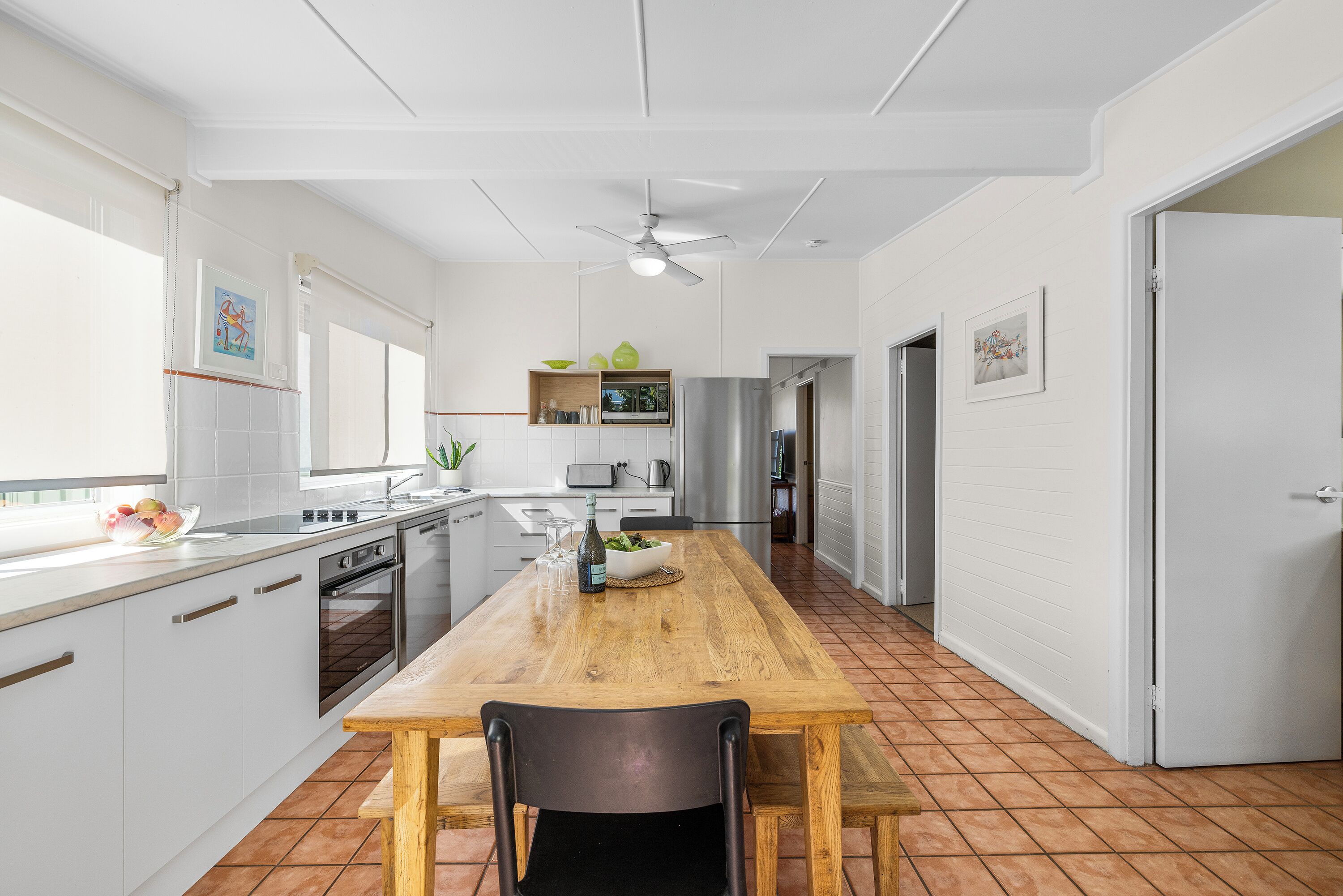 A modern kitchen with white cabinetry, a wooden dining table, and a ceiling fan.