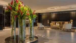 A modern lobby with a central round table surrounded by tall glass vases with red flowers, a dark-toned sofa, and a minimalist design. - LS Hotel (João Pessoa)