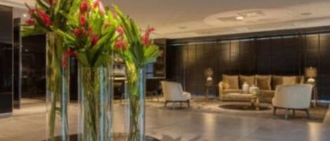 Een moderne lobby met een ronde centrale tafel, omringd door hoge glazen vazen met rode bloemen, een donkere bank en een minimalistisch design.