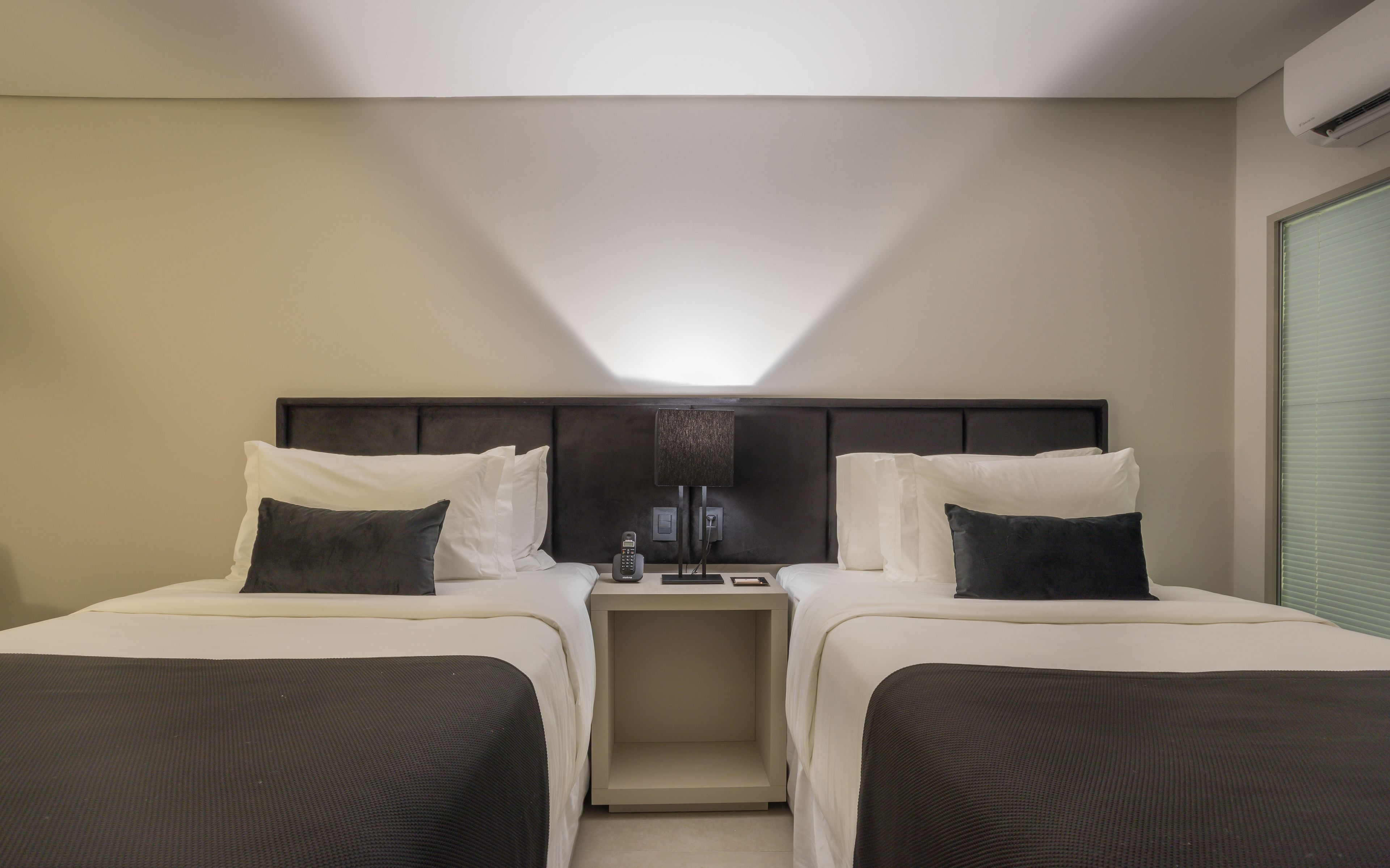 A hotel room with two beds, a bedside table with a lamp, and an air conditioning unit.