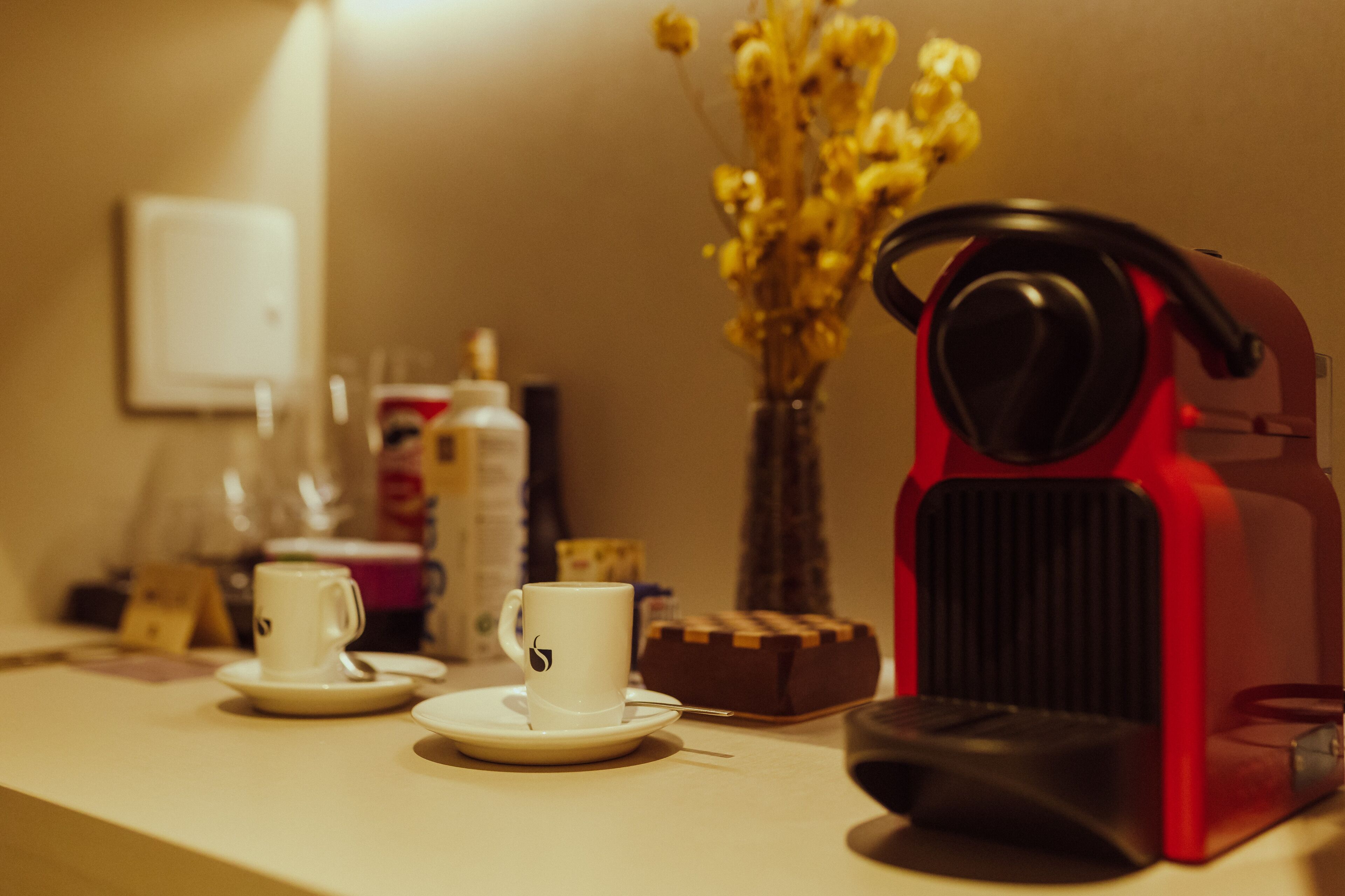 A coffee machine, two cups, a vase with yellow flowers, and a small wooden box on a table.