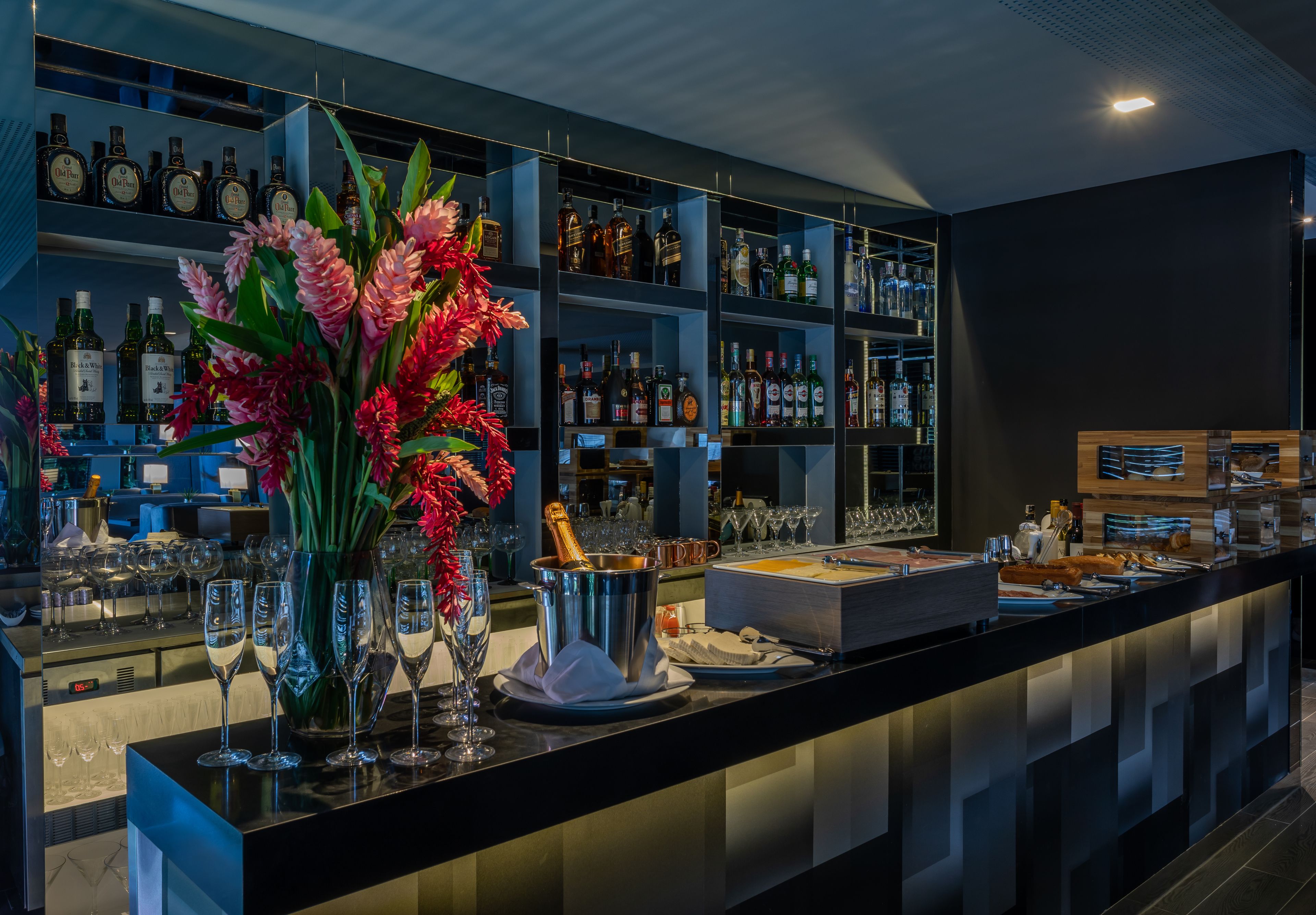 Zona de bar con estantes llenos de botellas de licor, un gran arreglo floral y una barra con vasos y un cubo para champán.