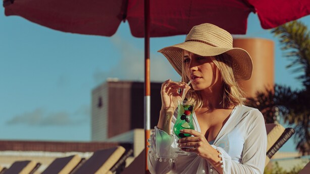 A person sitting on a lounge chair under a red umbrella, holding a drink. - LS Hotel (João Pessoa)