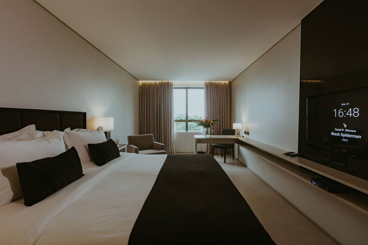 A hotel room with a bed, bedside tables, a desk, a chair, a TV, and a window with curtains.