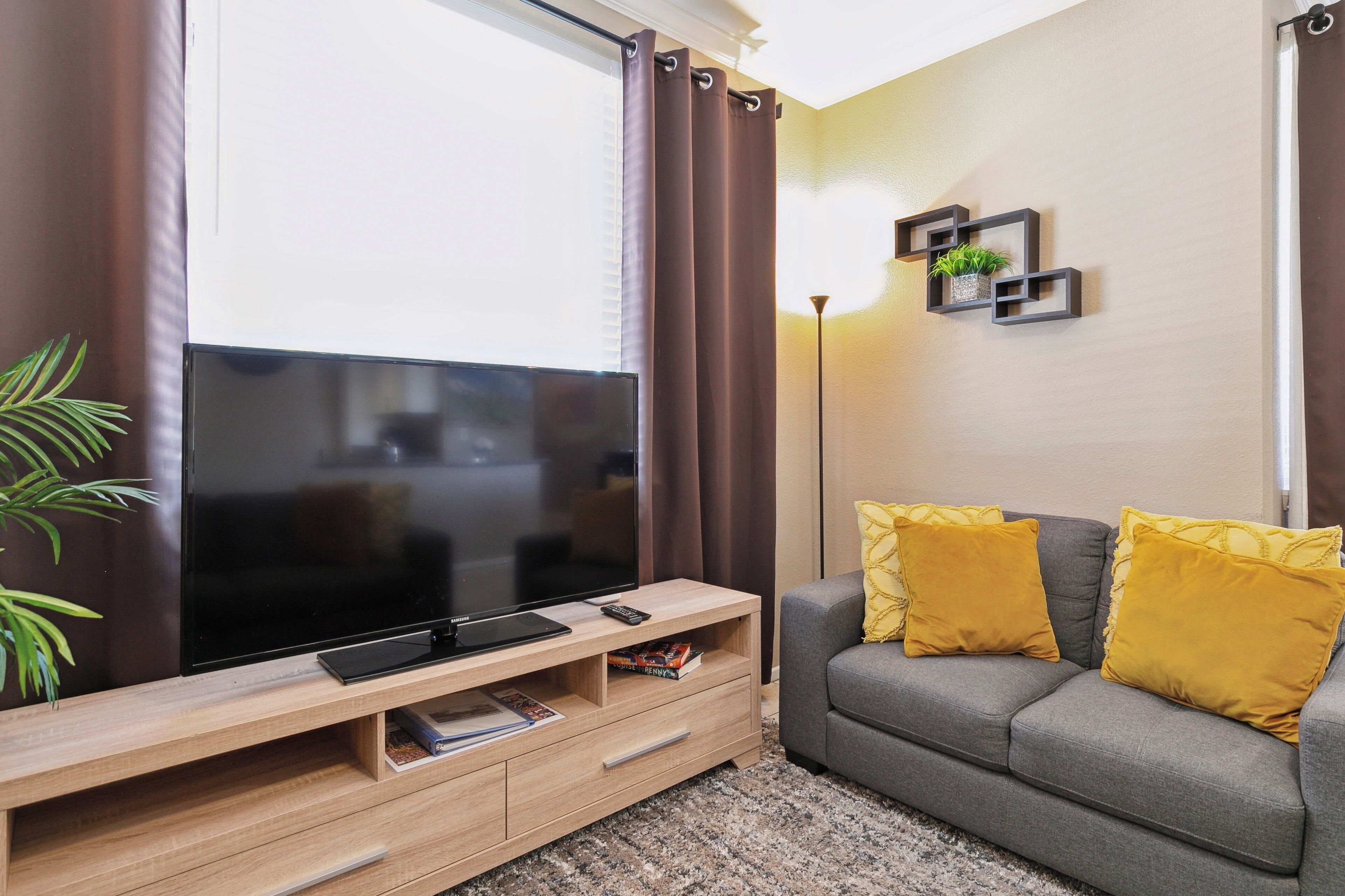 A living room with a flat-screen TV on a wooden TV stand, a grey sofa with yellow cushions, and a plant on the left.