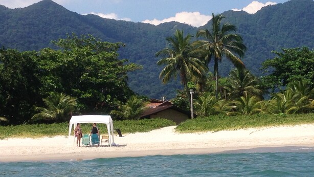 A beach with a white canopy and palm trees, a hut in the background, and mountains in the distance. - Pleasant beachfront house on Guaecá beach, São Sebastião, SP (São Sebastião)