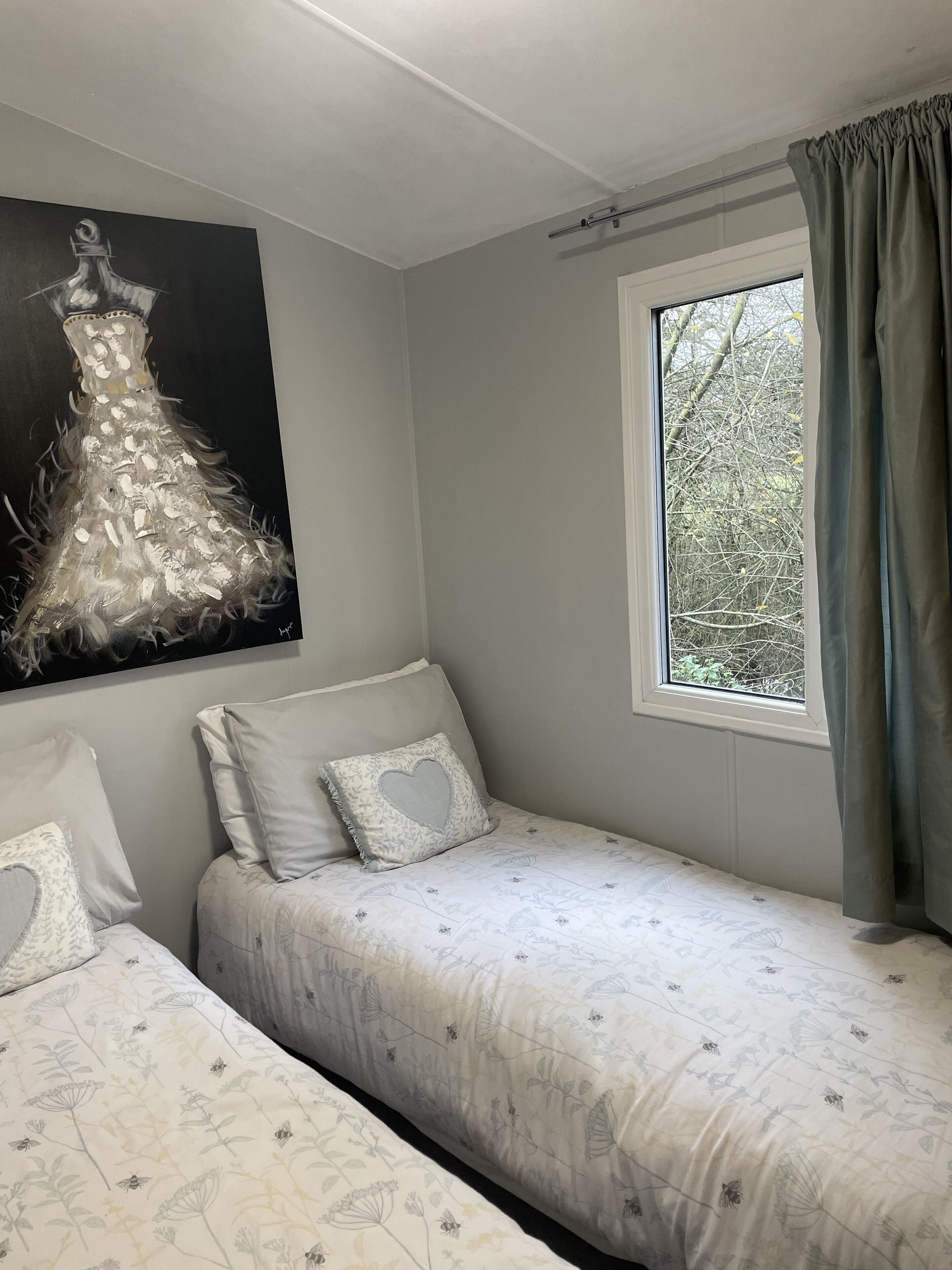 Ein Schlafzimmer mit zwei Einzelbetten, einem Fenster mit Vorhängen, einem Bild an der Wand und Blick auf Bäume.
