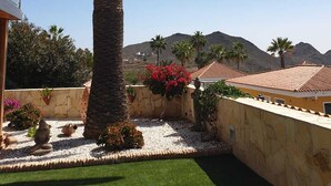 A palm tree in a landscaped area with a stone wall, a small garden with red flowers, and a building with a tiled roof in the background. - Casa Alma 
Beautiful Villa with Mountain & Sea Views
3 Bedrooms 2 Bathrooms (Chayofa)
