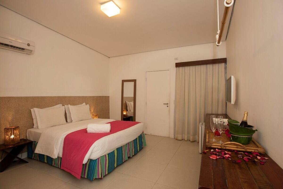 A hotel room with a bed, bedside table, air conditioning unit, curtains, and a minibar with glasses and a bottle.