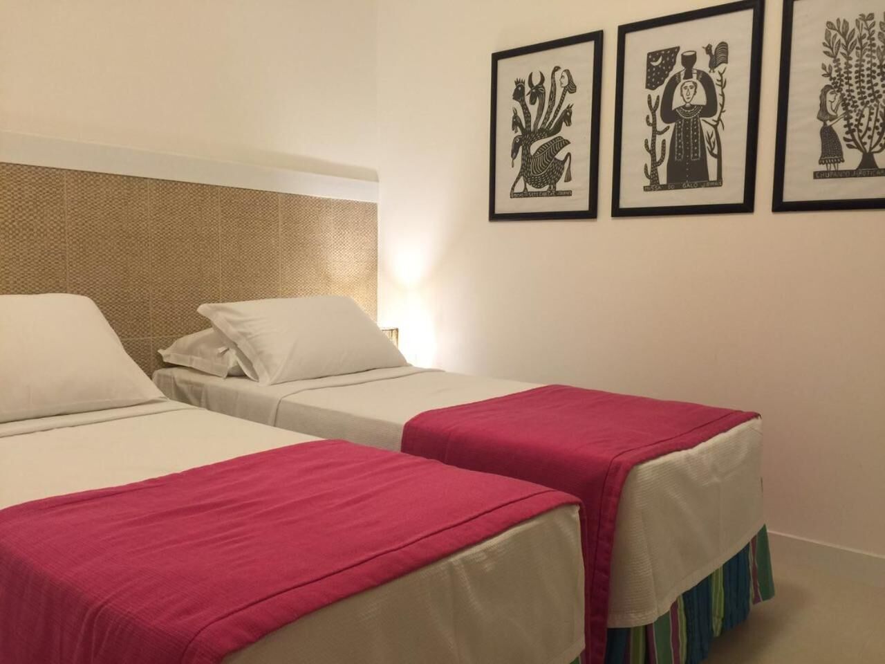 Een hotelkamer met twee bedden, elk met een rode deken, witte kussens en een beige hoofdeinde. Er hangen drie kunstwerken in lijsten aan de muur.