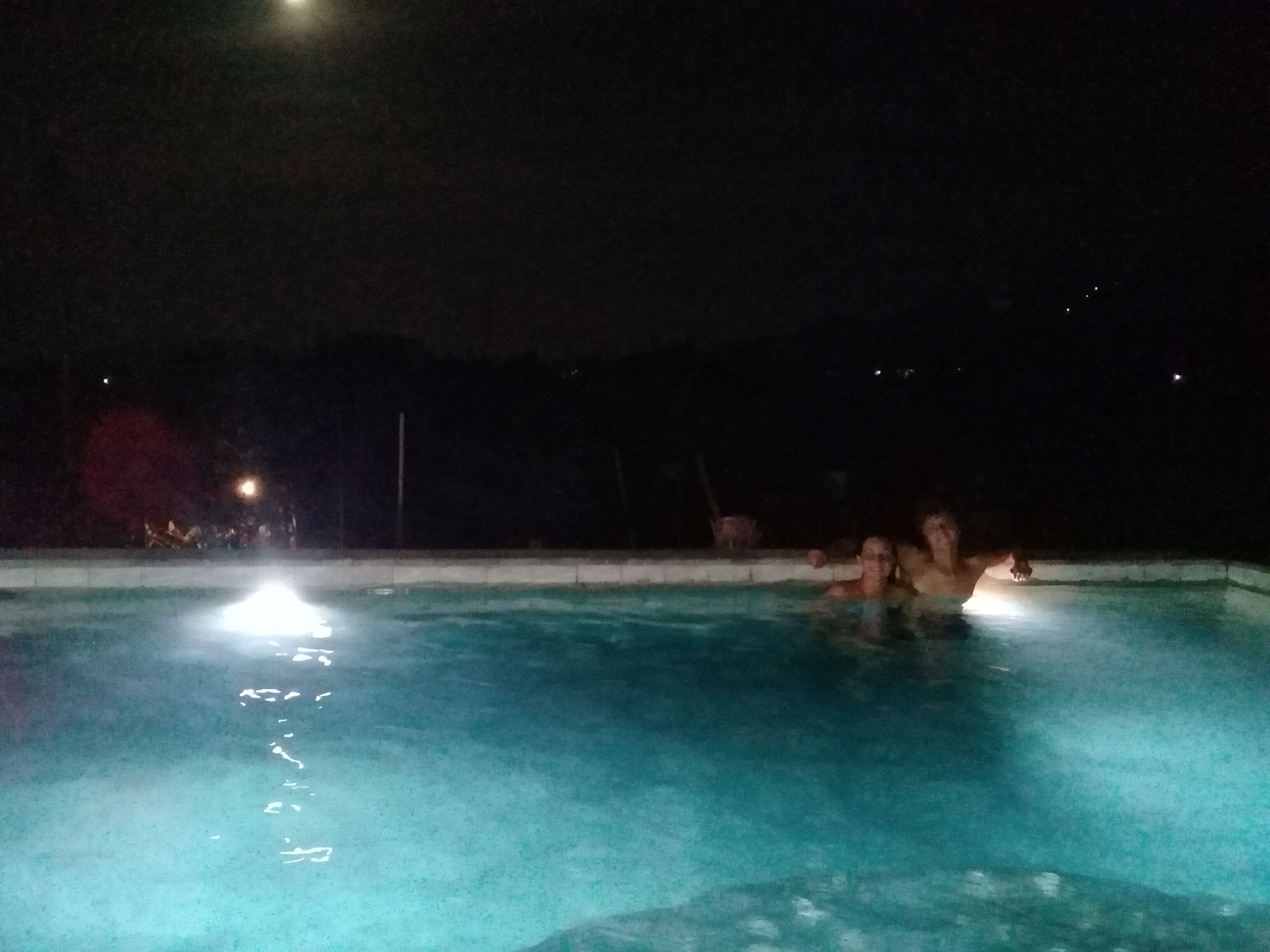 Piscina noturna com água cristalina e algumas pessoas na água.