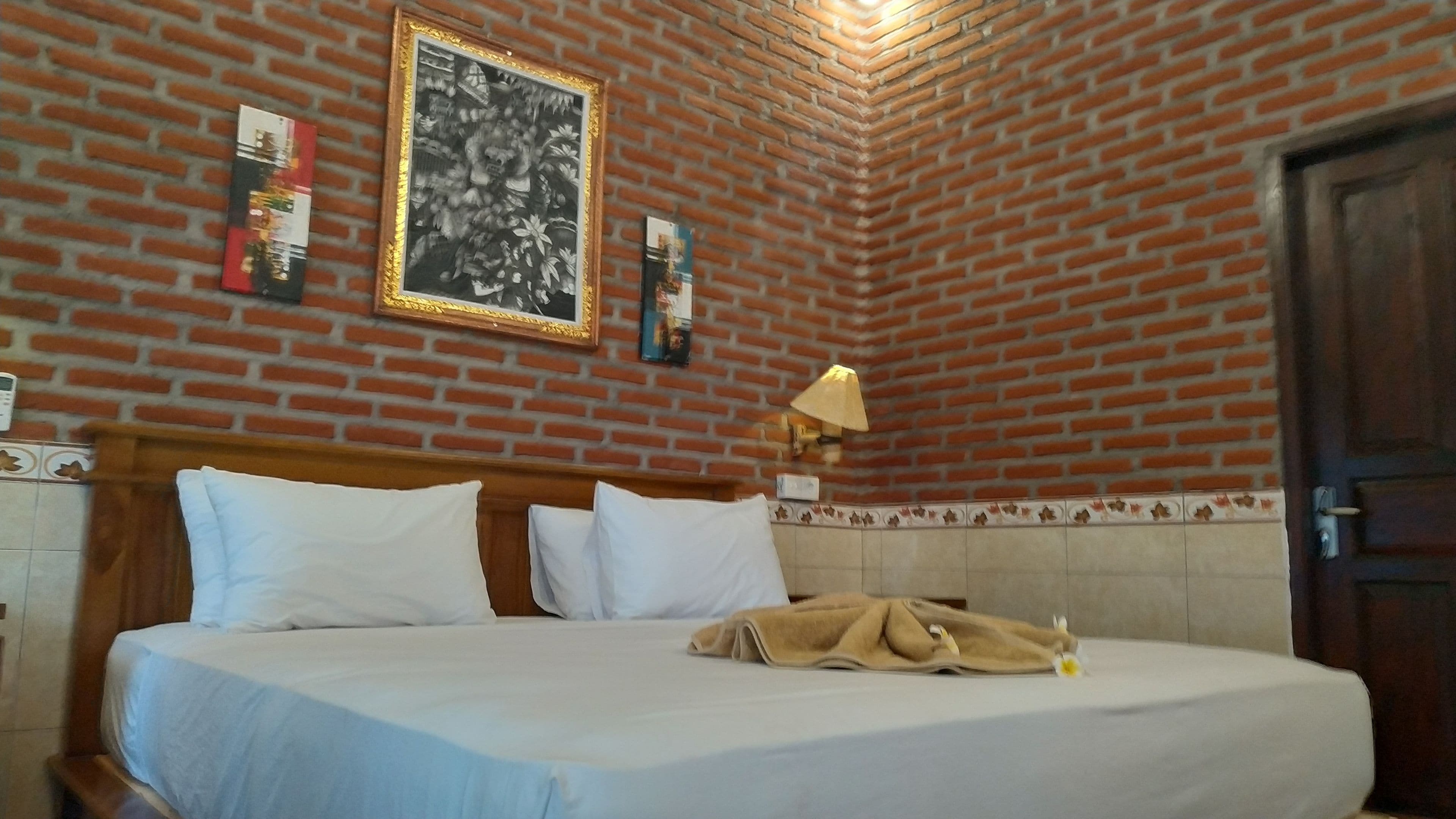 A hotel room with a brick wall, a bed with white bedding, a wooden headboard, a nightstand with a lamp, and a door.