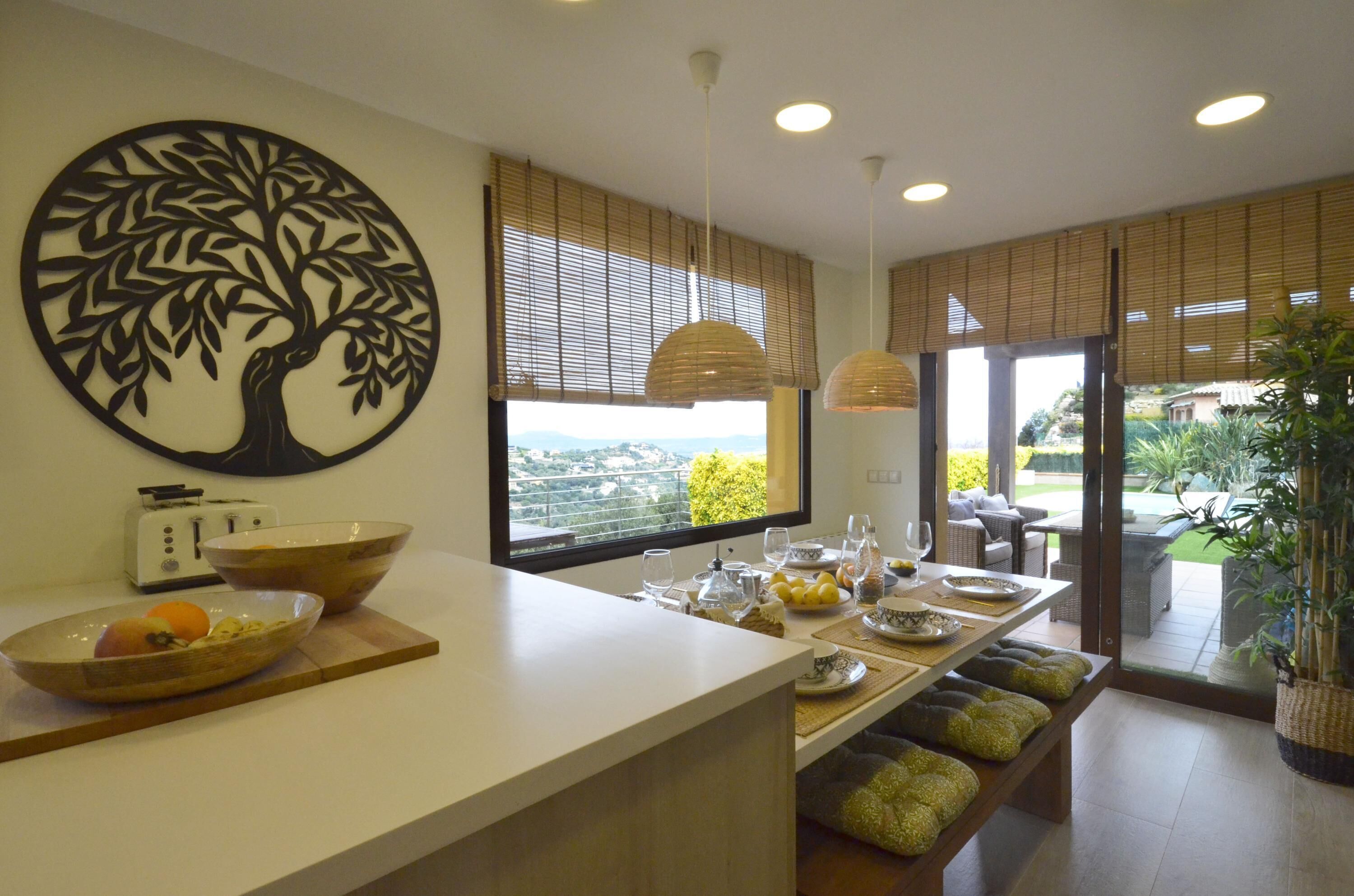 A modern kitchen with a large island, a dining area with a view, and a decorative tree wall art piece.