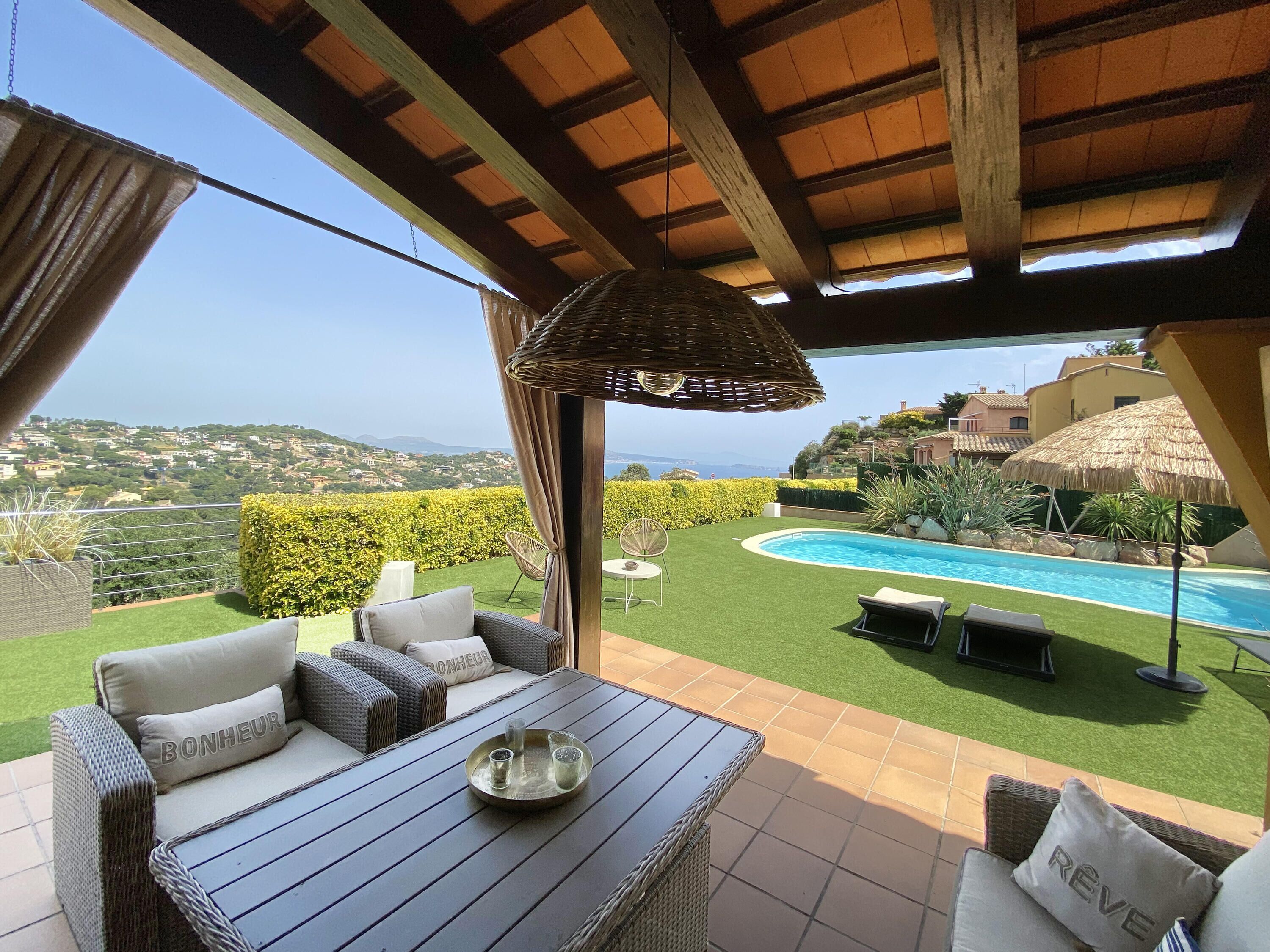 Eine Terrasse mit Korbmöbeln, einem Glastisch und Blick auf einen Swimmingpool und die Landschaft.