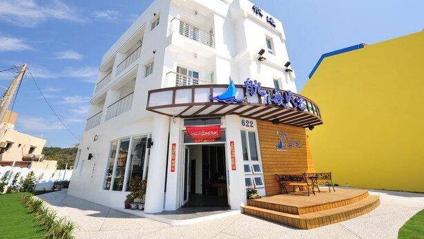 A white building with a blue sign, a wooden bench, and a red banner. - Hang Hai B & B (Hengchun)
