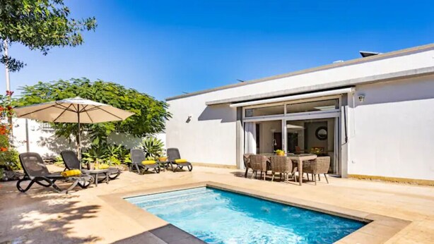 A swimming pool with lounge chairs and umbrellas, a dining area with a table and chairs, and a white building with a sliding glass door.