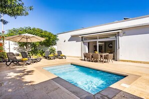 A swimming pool with lounge chairs and umbrellas, a dining area with a table and chairs, and a white building with a sliding glass door.