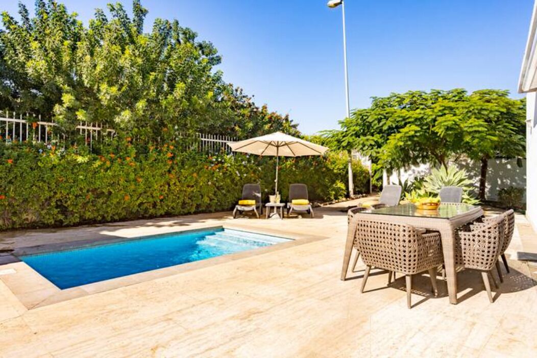 A swimming pool with lounge chairs and a table, surrounded by lush greenery and a white fence.