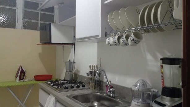 A kitchen with a gas stove, sink, and a blender. There are utensils hanging above the sink and a dish rack with plates and cups. - Apartamento Amoblado El Dorado (Bogotá)