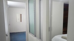 A hotel room with a bed, a window with blinds, a door, and a hallway leading to another room. - Apartamento Amoblado El Dorado (Bogotá)