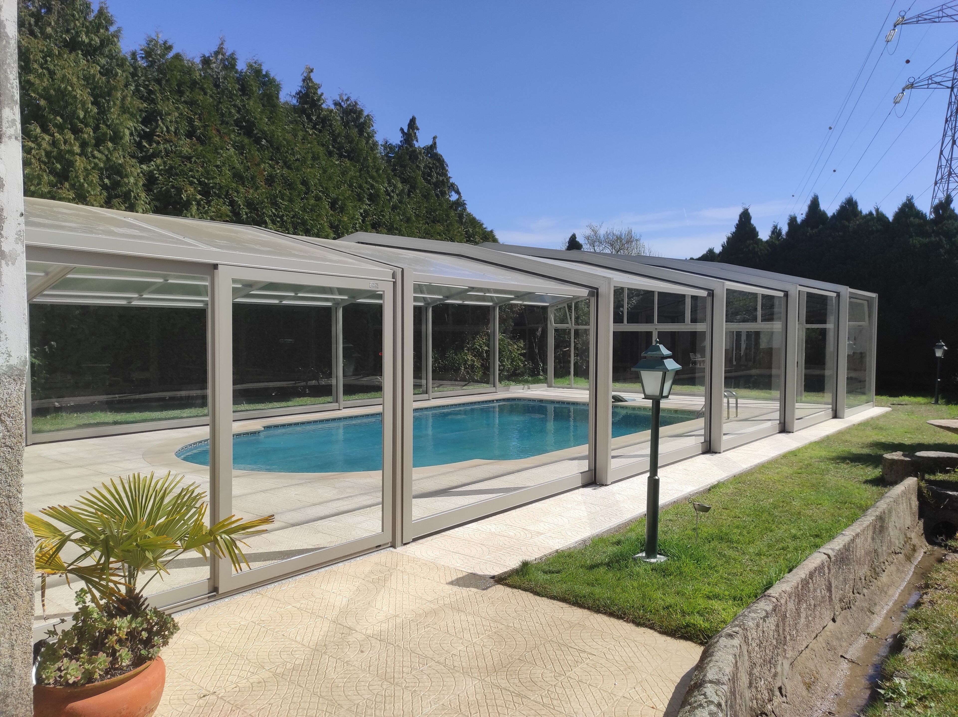 A covered swimming pool with a glass enclosure, surrounded by a paved patio and a grassy area.