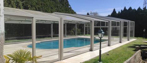 A covered swimming pool with a glass enclosure, surrounded by a paved patio and a grassy area.