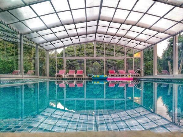 A covered swimming pool with clear glass walls, red lounge chairs, and a tiled floor.