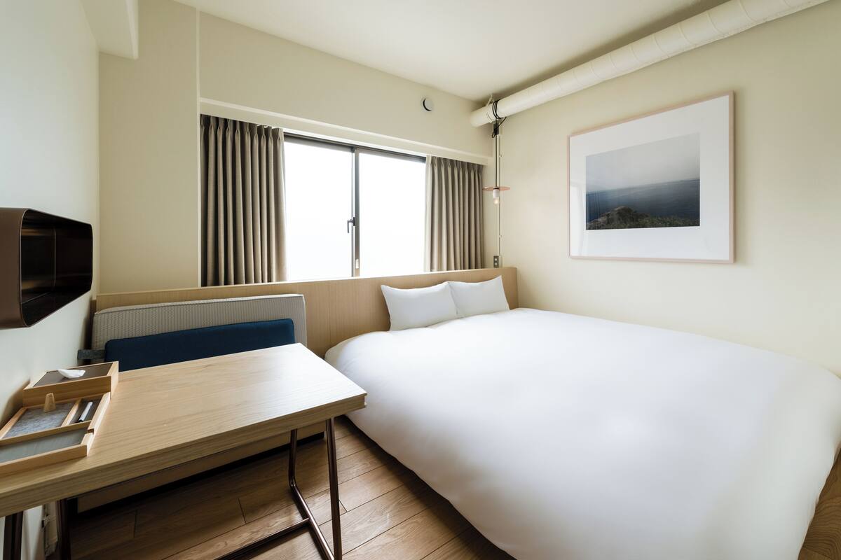 Una habitación de hotel con cama, escritorio, silla, radiador, ventana con cortinas e imagen enmarcada en la pared.