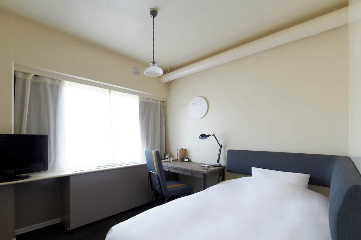 Habitación de hotel con cama, escritorio con ordenador, silla, lámpara de escritorio y ventana con cortinas.