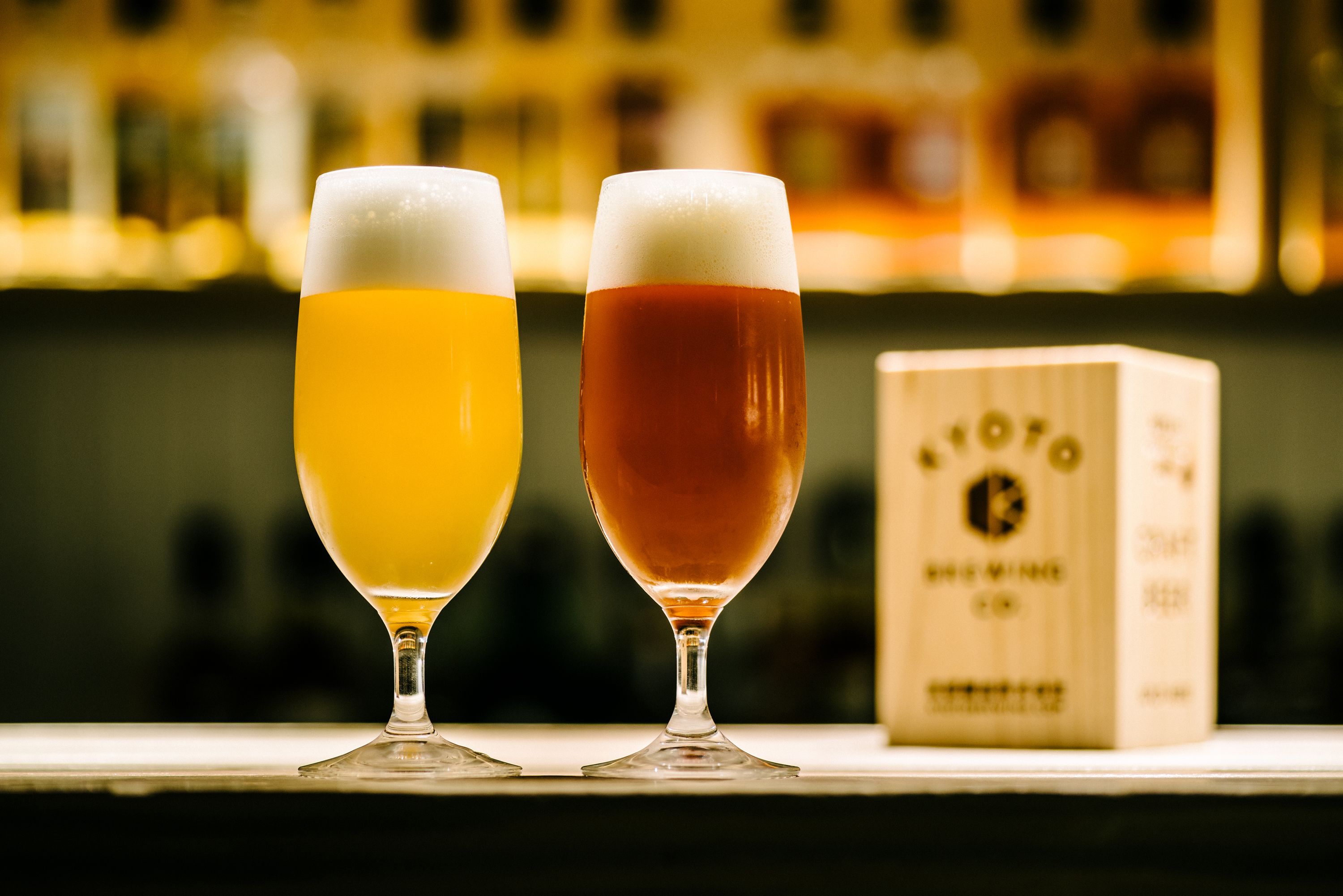 Two glasses of beer on a bar counter with a wooden box in the background.