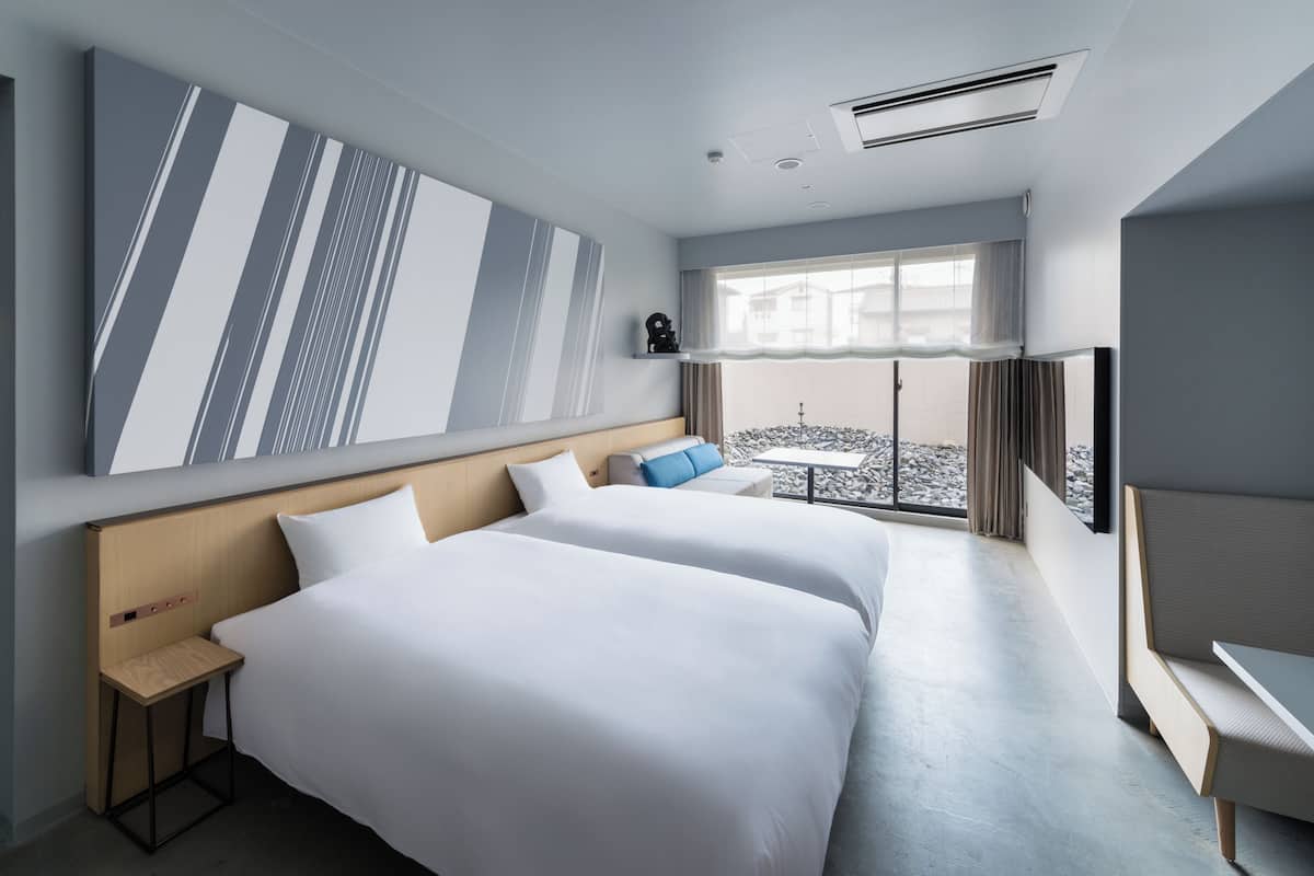 Habitación de hotel con dos camas, un ventanal amplio, vistas a edificios y un diseño moderno.