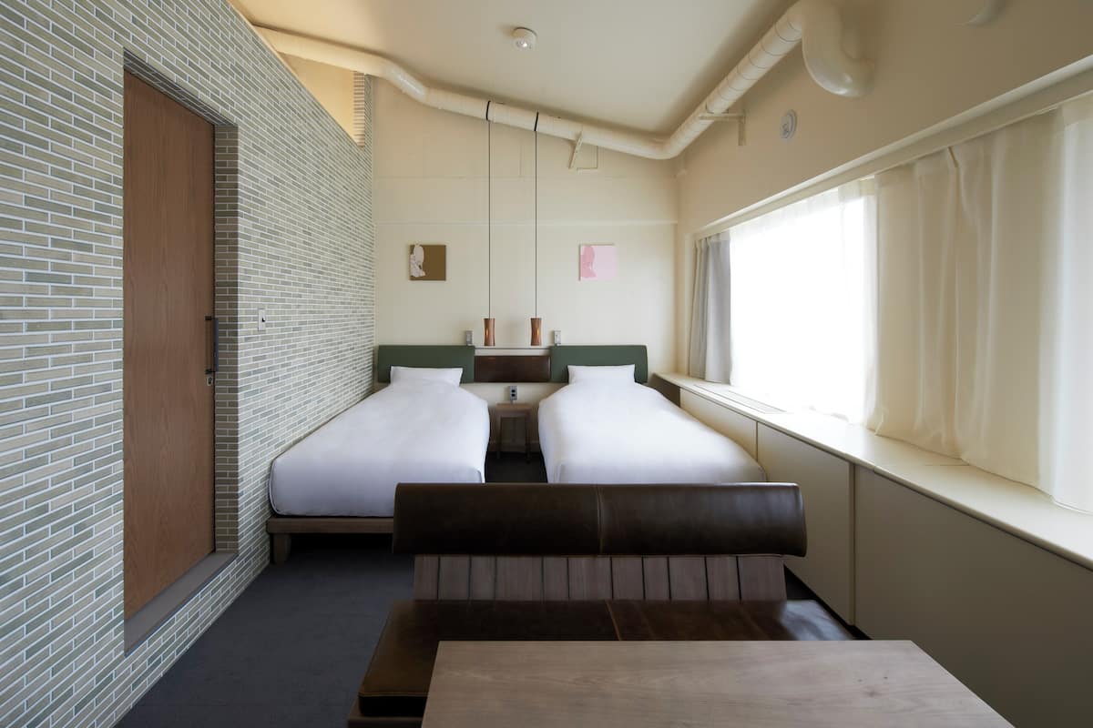 Habitación con dos camas, un banco, una mesa, una ventana con cortinas, pared de ladrillo y lámpara en el techo.
