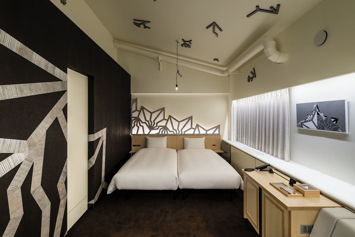 Una cama doble con un diseño geométrico en la pared, un escritorio con una lámpara y una ventana con cortinas.
