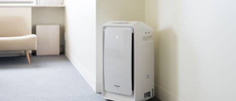A white air purifier in a corner of a room with a grey carpet and a white chair.