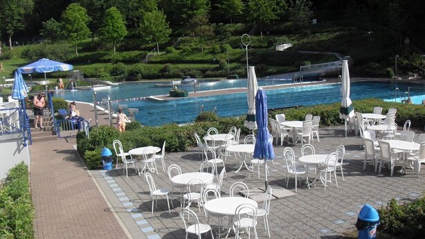 Zona de piscina al aire libre con varias mesas y sillas, rodeada de follaje y una piscina cristalina.