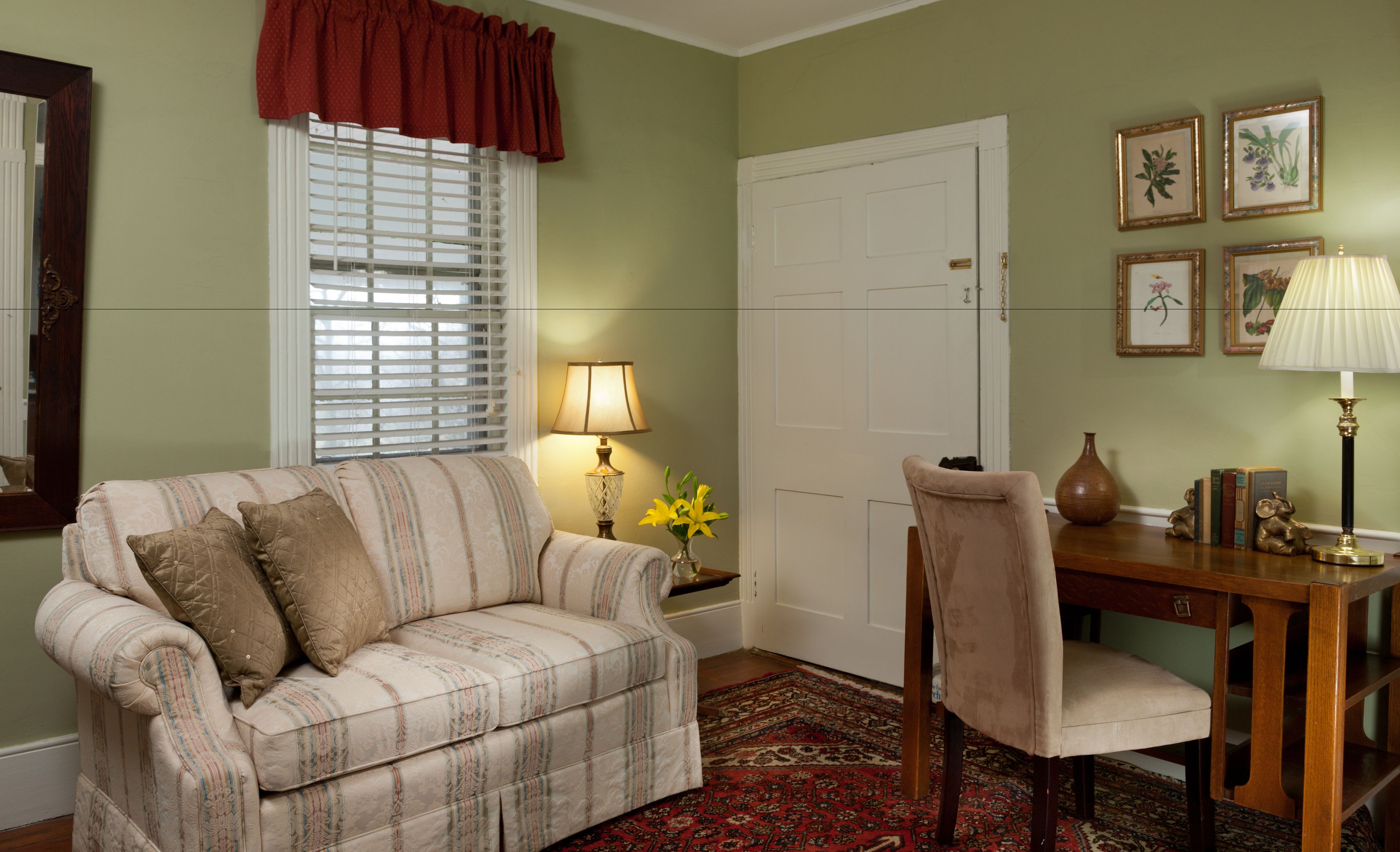 A room with a striped sofa, a wooden desk, a vase with flowers, and framed botanical prints on the wall.