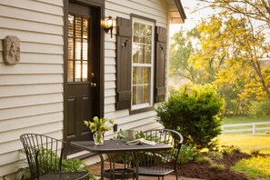 A wooden house with a front door, two windows with shutters, a small garden with a white fence, and a table with chairs outside.