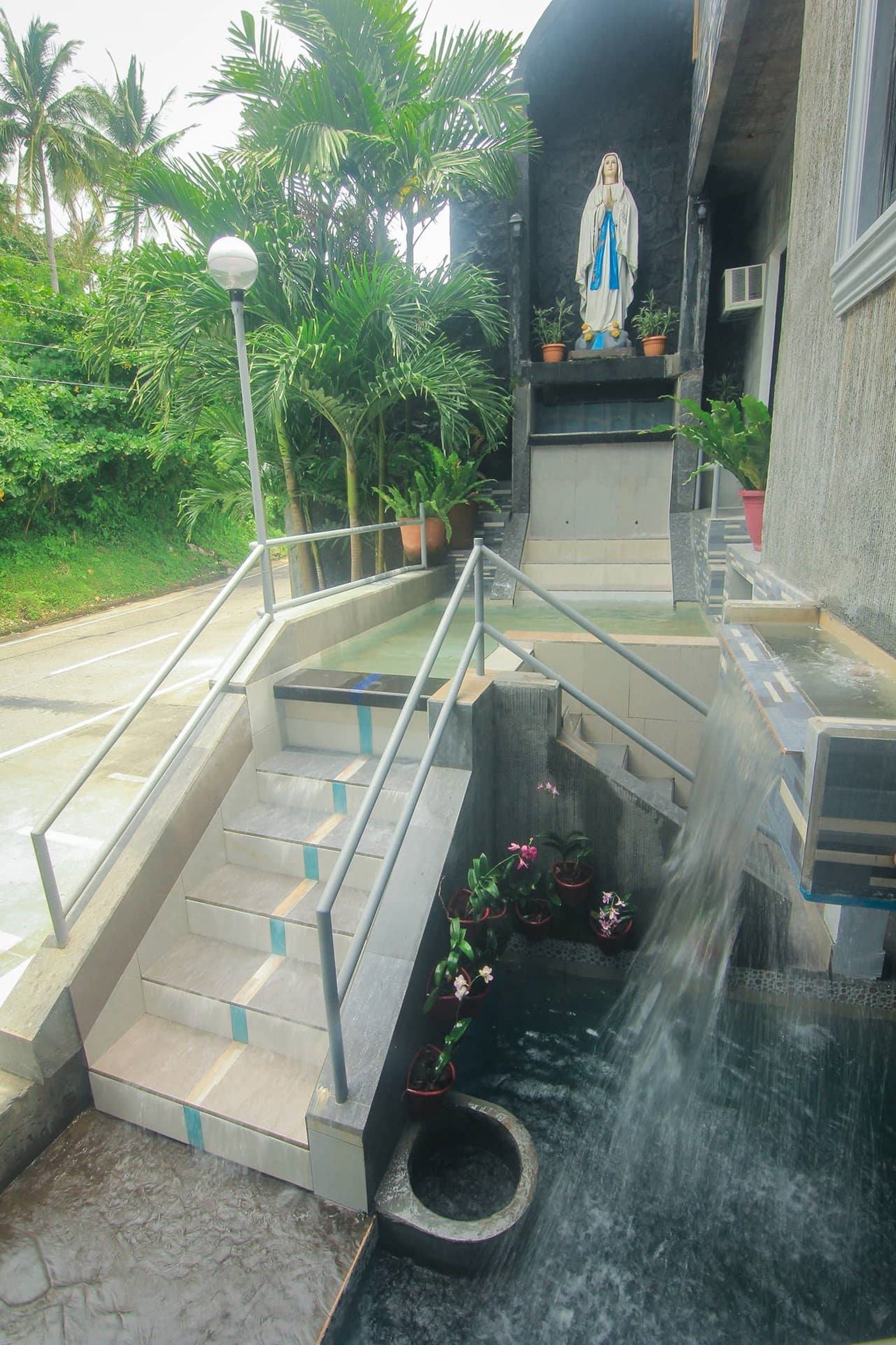 Una estatua sobre un pedestal con escaleras que la conducen, rodeada de plantas en macetas y una fuente.