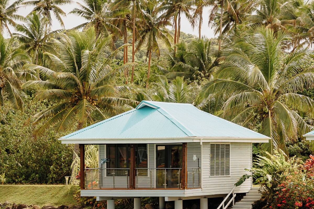 A two-story house with a blue roof, situated on stilts over a body of water, surrounded by palm trees.