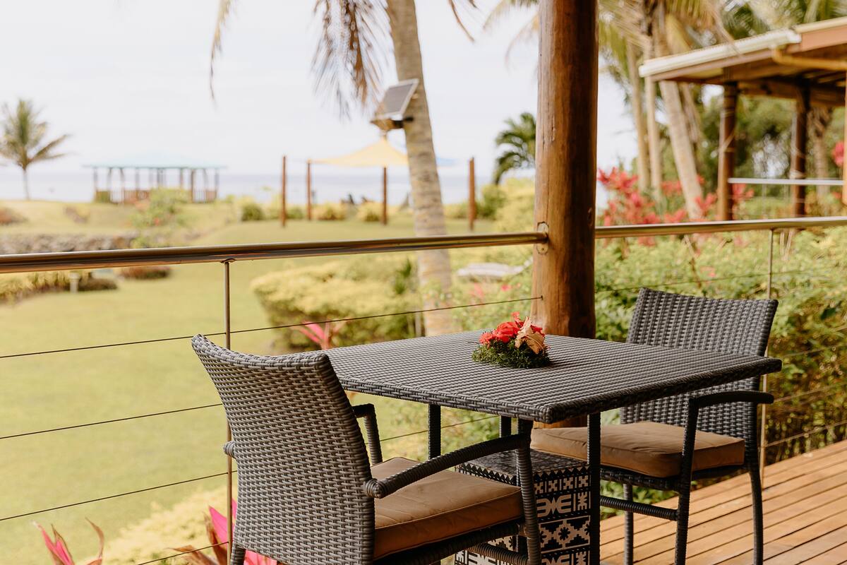 A wicker table with a cushion and a small flower arrangement on a wooden deck overlooking a landscaped garden with a view of the ocean.