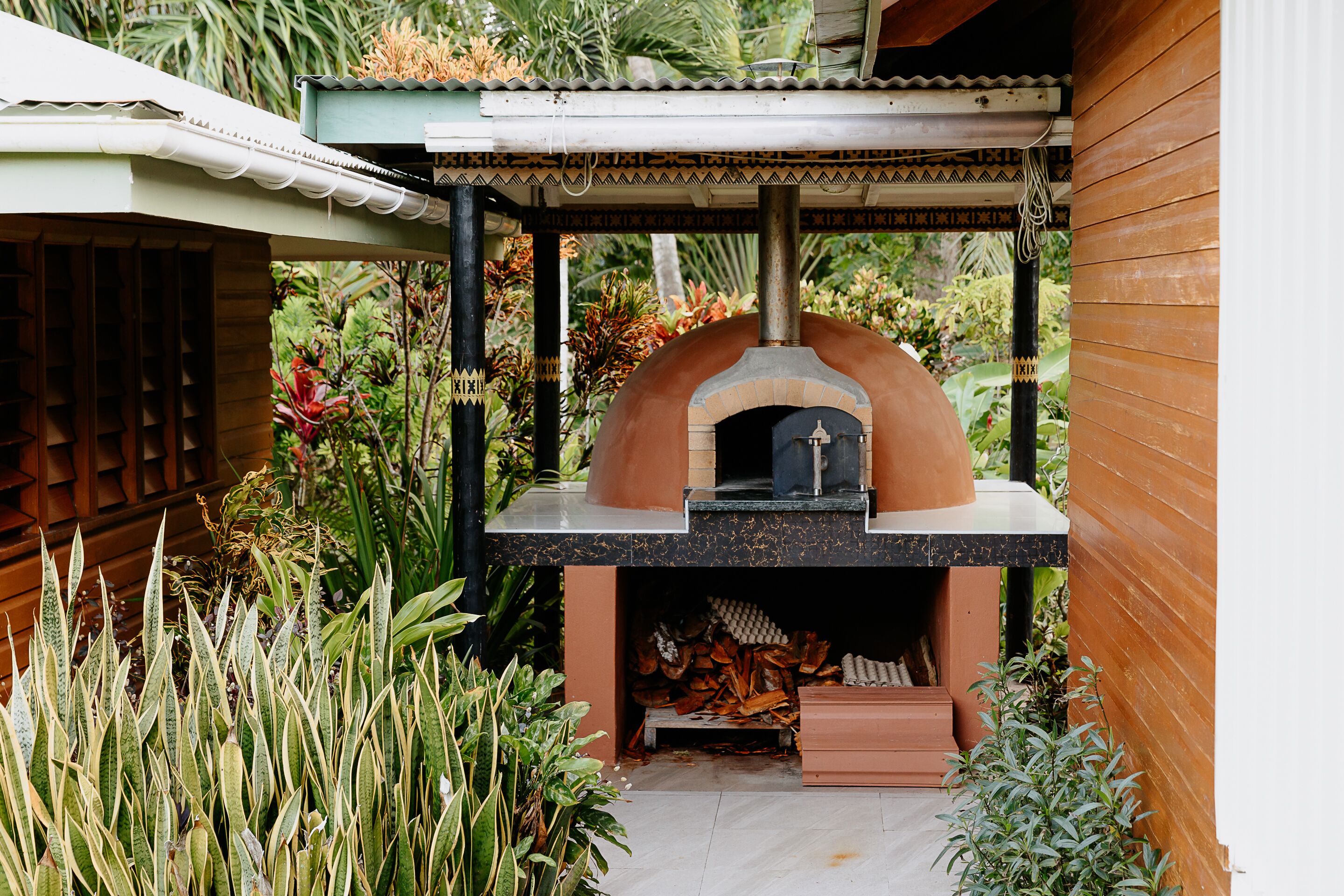 An outdoor brick oven with a chimney, surrounded by lush greenery and a wooden structure.