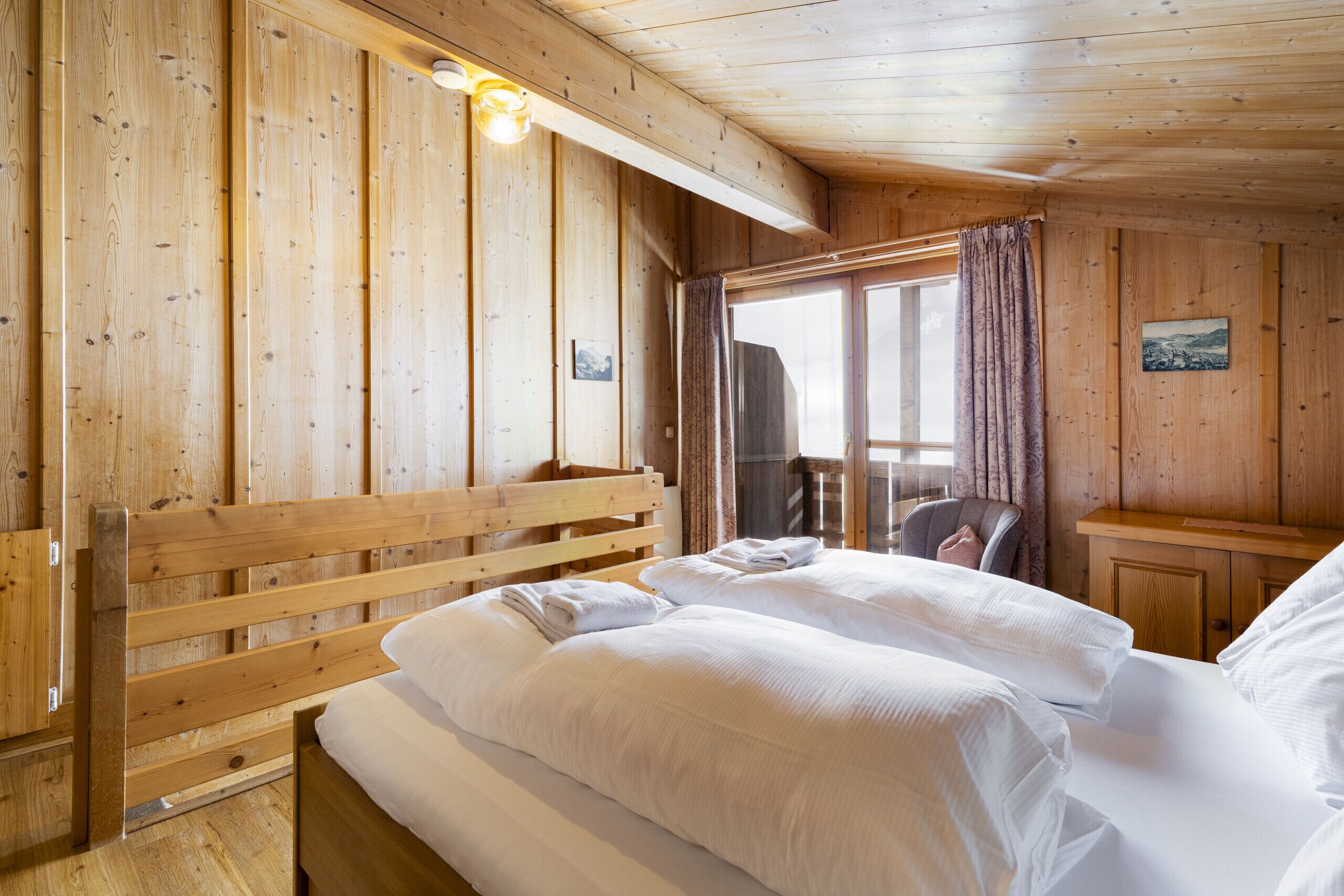 A wooden cabin room with a bed, a chair, and a window with curtains.